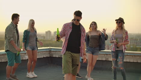 Young-man-is-dancing-with-beer-on-the-roof-with-girls-and-boys-friends.-It's-a-hot-summer-evening-on-the-the-roof.-He-is-wearing-short-green-shorts-a-red-plaid-shirts-and-fashionable-glasses.